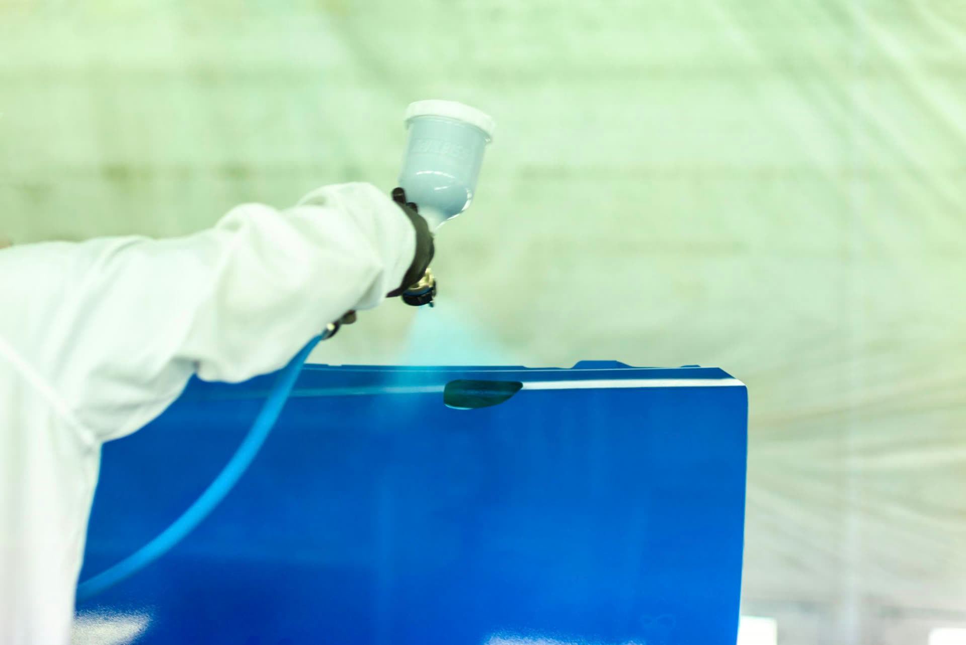 Painter in protective suit applying base coat to a blue panel inside the spray booth